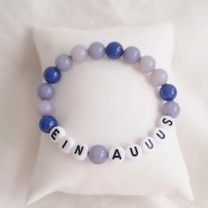 Peut inclure: Un bracelet de perles alternant des perles bleues et lavande. Le bracelet comporte des perles de lettres blanches épelant "EIN AUUS". Le bracelet est présenté sur un coussin blanc.