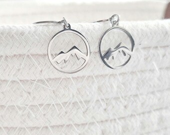 May include: A pair of silver-colored earrings. Each earring features a mountain range design within a circular frame. The earrings are displayed against a white, textured background.