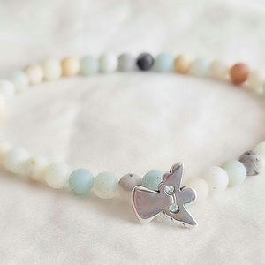 Puede incluir: Una pulsera de cuentas pequeñas, redondas y mate en tonos suaves de azul pálido, crema, beige y gris. Un dije de metal plateado pulido con forma de ángel estilizado con las alas extendidas está sujeto a la hilera de cuentas.