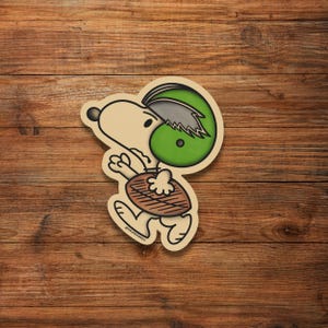 May include: A cartoon illustration of Snoopy wearing a green and black football helmet and holding a football. The background is a wood grain pattern.