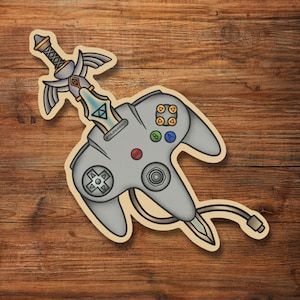 May include: A gray Nintendo 64 controller with a gold Master Sword from The Legend of Zelda sticking out of the top. The controller has a tan background and the sword has a blue and silver blade.