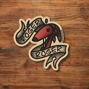 May include: A sticker featuring a red ram's head with the word "ROGER" in a banner above and below. The design is in a traditional tattoo style, with black outlines and a cream-colored background. The sticker is on a wooden surface.