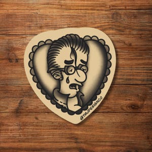 May include: A tattoo-style sticker featuring a crying cartoon character with glasses, set against a heart-shaped background. The design is in black and gray tones, with the text "@TMJDESIGNCO" visible.