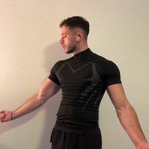 Könnte beinhalten: Schwarzes, sportliches Kompressionsshirt mit kurzen Ärmeln und Stehkragen. Das Shirt hat ein dezentes, strukturiertes Design auf der Brust und an den Seiten. Die Person trägt auch schwarze Sporthosen.