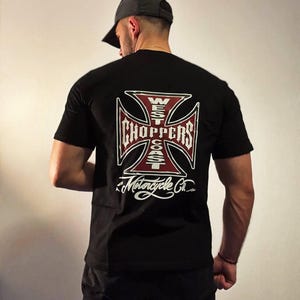 May include: Black t-shirt with a large, detailed graphic on the back. The graphic is a stylized cross with the words "WEST COAST CHOPPERS" and "Motorcycle Co." in white and red. The shirt is short-sleeved.