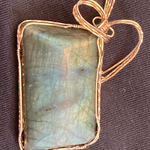 May include: A rectangular pendant featuring a large, iridescent, blue-green stone. The stone is encased in a gold-colored wire frame with a looped design at the top for hanging. The pendant has a handmade, artisan look.