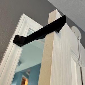 May include: Black metal door stop with a triangular shape, mounted on a white door frame.