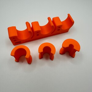 May include: Orange plastic pipe holder with three slots and three additional pieces. The holder is designed to hold pipes in place.