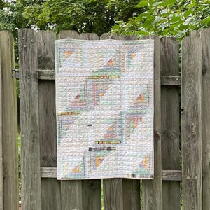 Peut inclure: Une courtepointe blanche avec un motif de patchwork composé de carrés de différentes couleurs et motifs. La courtepointe est accrochée à une clôture en bois.