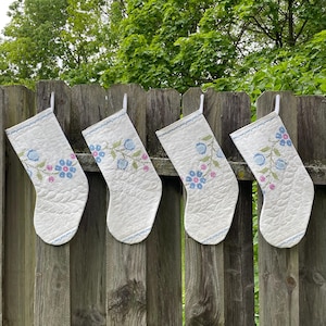 May include: Four white Christmas stockings with floral embroidery in blue and pink hang on a wooden fence. The stockings have a quilted pattern and a blue trim along the top edge. The background features green foliage.