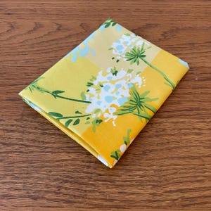 Puede incluir: Una tela amarilla con un patr&oacute;n floral blanco. Las flores son estilizadas y tienen un centro azul claro. La tela est&aacute; doblada por la mitad.