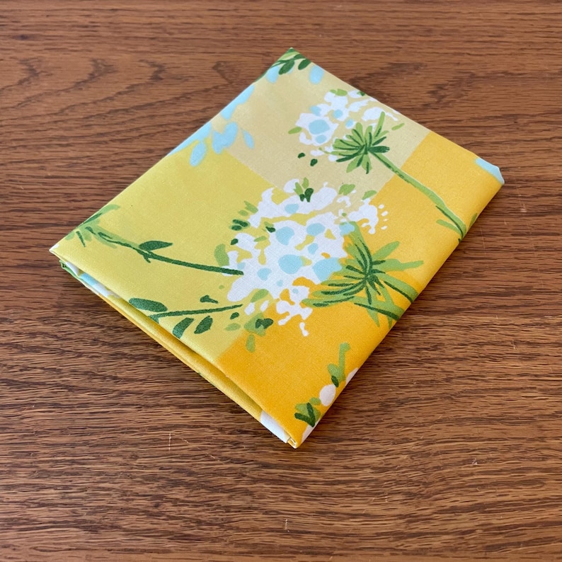 Puede incluir: Una tela amarilla con un patr&oacute;n floral blanco. Las flores son estilizadas y tienen un centro azul claro. La tela est&aacute; doblada por la mitad.