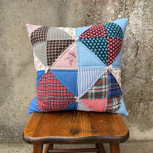 May include: Square patchwork pillow with a variety of fabric patterns, including plaid, stripes, and floral designs. The pillow features a mix of colors, including blue, red, pink, and brown. The pillow is resting on a wooden stool.