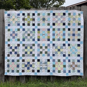 May include: A patchwork quilt featuring a grid of square blocks in various shades of blue, green, pink, and white. The design consists of smaller squares arranged in a nine-patch pattern, separated by light blue sashing and white borders.