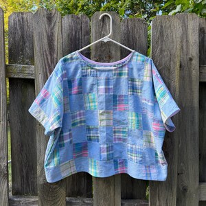 May include: A short-sleeved top featuring a patchwork design of light blue solid fabric squares alternating with squares of blue, pink, green, and white plaid fabric. The garment has a wide, boxy fit, a round neckline, and a light purple interior trim.