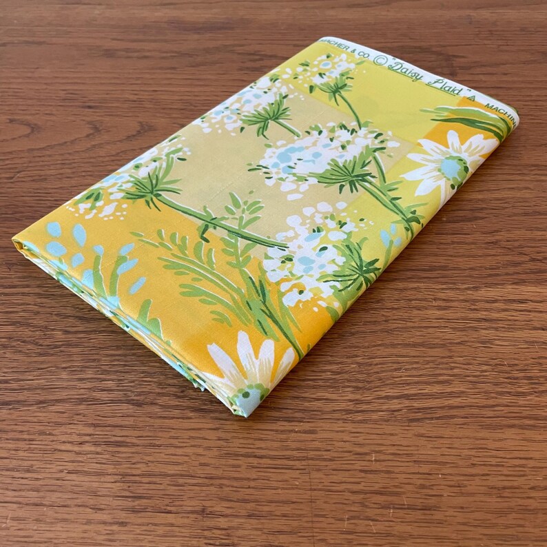 Puede incluir: Una tela amarilla con un patr&oacute;n de margaritas blancas y hojas verdes. La tela tiene el texto "Archer & Co. Daisy Plaid" impreso en ella.