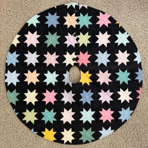 May include: A circular quilted tree skirt featuring a black background with a repeating pattern of eight-pointed stars in pastel shades of pink, blue, yellow, green, and cream. The skirt has a central opening and a slit with three black buttons.