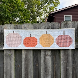 May include: A rectangular quilted table runner featuring four stylized pumpkin shapes in shades of light pink, orange, yellow, and dark red. Each pumpkin has a small brown stem and a grid-like fabric pattern, set against a white background.