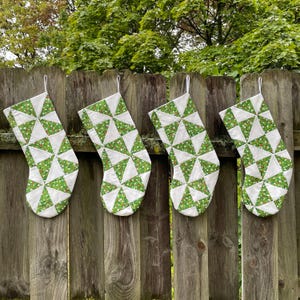 Peut inclure: Quatre bas de Noël sont suspendus à une clôture en bois. Chaque bas présente un fond blanc avec des triangles verts et de petits accents floraux rouges. Les bas sont en tissu et ont une boucle blanche pour les suspendre.