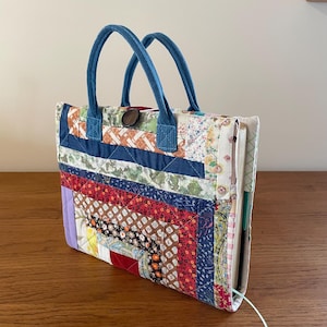 May include: A handmade patchwork tote bag with blue denim handles. The bag features a variety of fabric patterns in red, blue, brown, and floral prints. A wooden button secures the top. The bag is designed to hold a notebook or journal.