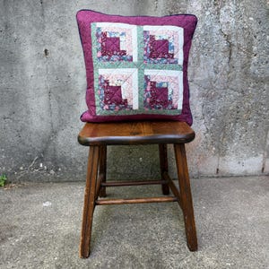May include: A quilted pillow with a patchwork design in shades of purple, pink, and green, resting on a wooden stool. The pillow features a square pattern with a dark purple border and a navy blue trim.