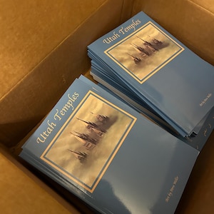 Puede incluir: Una pila de libros de bolsillo azules con el título "Utah Temples" y las palabras "Art by Steve Miller" en la portada. Los libros están en una caja de cartón.