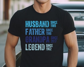 May include: A black short-sleeved t-shirt featuring blue text that reads: HUSBAND SINCE 1985, FATHER SINCE 1986, GRANDPA SINCE 2017, and LEGEND SINCE 1962. The graphic design uses a distressed font style printed on the front of the garment.