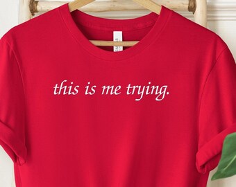May include: A bright red t-shirt with the phrase "this is me trying" printed in white, centered on the chest. The short-sleeved shirt is knotted at the waist and hangs on a wooden hanger. The background includes a wooden ladder and decorative plants.