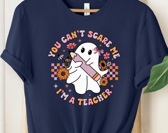 May include: Navy blue t-shirt featuring a ghost holding a pencil, surrounded by flowers and the text "YOU CAN'T SCARE ME I'M A TEACHER". The design includes a checkerboard pattern and a retro color palette. The shirt is displayed on a wooden hanger.