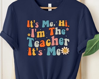 May include: A navy blue short-sleeve t-shirt featuring the multi-colored text It's Me, Hi, I'm The Teacher It's Me in a retro font. The design includes small colorful flower icons and a smiling daisy graphic on the front of the garment.