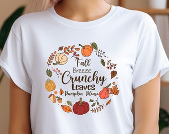 May include: A white short-sleeved t-shirt featuring a circular graphic of autumn elements including pumpkins, leaves, and berries. The text in the center reads Fall Breeze Crunchy Leaves Pumpkin Please in a mix of script and block lettering.