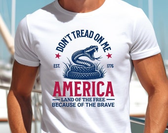 May include: White t-shirt with a graphic design featuring a coiled blue snake, the text "DON'T TREAD ON ME," and the word "AMERICA." The design includes the text "LAND OF THE FREE BECAUSE OF THE BRAVE" and the year "1776."