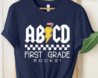 May include: Navy blue t-shirt with the text "ABCD FIRST GRADE ROCKS!" in a white, rock-and-roll style font. The design includes a yellow lightning bolt with a pink and yellow pencil. The shirt is hanging on a wooden hanger.