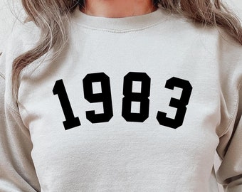 May include: A light gray sweatshirt with black block lettering that reads "1983".