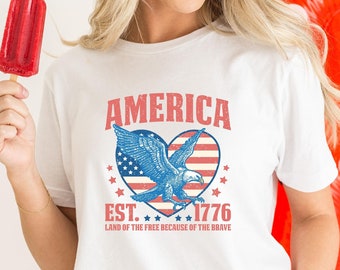 May include: White t-shirt with a patriotic design featuring the word "AMERICA" above a heart-shaped graphic of an eagle and the American flag. The text "EST. 1776 LAND OF THE FREE BECAUSE OF THE BRAVE" is below the graphic.