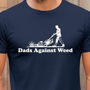 May include: Navy blue t-shirt with a white graphic of a person mowing a lawn, and the text "Dads Against Weed". The shirt is made of a soft material and has short sleeves.