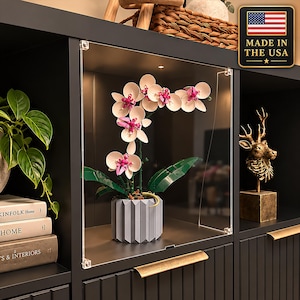 May include: A clear acrylic display case housing a buildable orchid plant with light pink petals, dark pink centers, and green leaves in a gray fluted pot. The case features metal corner connectors and is positioned on a black shelf unit.