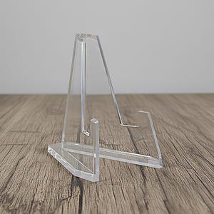 May include: A clear acrylic display stand with a triangular support and a horizontal ledge. The stand is designed to hold and showcase items. The stand is made of clear acrylic and has a modern, minimalist design.