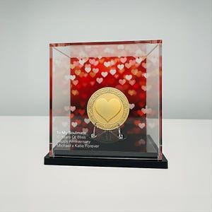 May include: A gold-colored heart-shaped medallion inside a clear acrylic display case. The medallion is on a stand, set against a red background with heart-shaped bokeh. Text on the case reads "To My Soulmate 10 Years Of Bliss Happy Anniversary Michael x Katie Forever."
