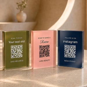 Può includere: Tre insegne rettangolari in acrilico nei colori verde oliva, rosa tenue e blu navy. Ogni insegna presenta un codice QR stampato e testo: 'SCAN TO BOOK Your next visit SAGE WELLNESS', 'SCAN TO LEAVE A Review LUNA BEAUTY' e 'FOLLOW US ON Instagram NOVA STUDIO'.