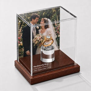 May include: A gold ring with a large center stone sits on a clear stand inside a glass display case with a dark wood base. The case features a photograph of a couple and the text: The Eternity Ring, Love Lasts Forever, 29th September 2016.