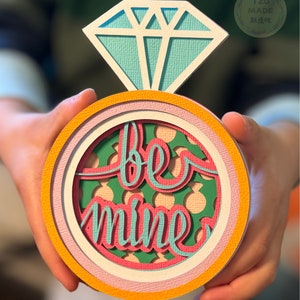 May include: A paper craft ring with a large blue diamond on top and the words "be mine" inside the ring. The ring is layered with pink, white, and yellow paper.