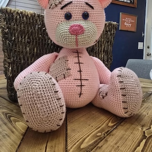May include: A handmade pink crochet teddy bear with a patched ear and chest. The bear has black stitched details, a tan muzzle, and blue eyes. The bear is sitting on a wooden surface.