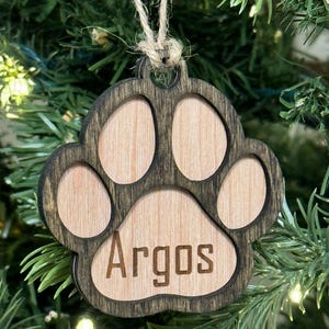 May include: Wooden paw print ornament with the name "Argos" engraved in the center. The ornament is made of light-colored wood with a dark brown border and a twine loop for hanging. The ornament is displayed on a Christmas tree.
