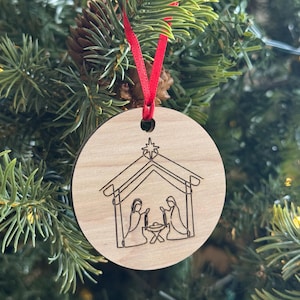 May include: A round wooden Christmas ornament with a laser-etched nativity scene. The ornament features a red ribbon for hanging and is displayed on a Christmas tree with green needles and a pine cone.