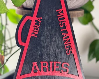 May include: A black and red wooden megaphone-shaped sign. The sign features the text "2025", "NBCA", "MUSTANGS", and "ARIES" in red. The sign is likely a school or team-related item.