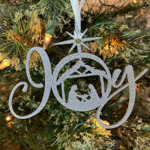 May include: A silver glitter Christmas ornament with the word "Joy" in a cursive font. The ornament features a nativity scene and a starburst design, hanging from a white ribbon. The ornament is displayed on a Christmas tree.