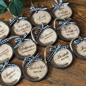 May include: Wooden Christmas ornaments with various religious phrases, including "King of Kings" and "Jesus." Each ornament has a dark brown border, a light wood center, and a black and white checkered bow. The ornaments are connected with twine.