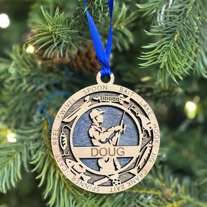 May include: A wooden Christmas ornament featuring a silhouette of a person fishing. The ornament is personalized with the name "DOUG" and includes various fishing terms around the edge, such as "reel", "cast", "bait", "spoon", "hook", and "jig".