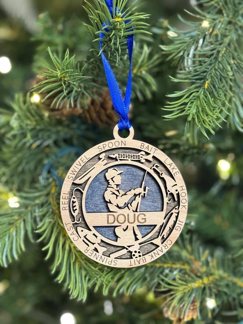 May include: A wooden Christmas ornament featuring a silhouette of a person fishing. The ornament is personalized with the name "DOUG" and includes various fishing terms around the edge, such as "reel", "cast", "bait", "spoon", "hook", and "jig".